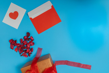 a red heart on a white paper, a red envelope, dried red leave and brown gift box tied with red ribbon put on the left of the picture on a blue background