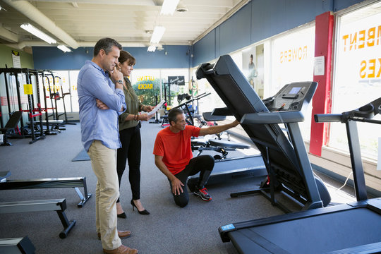 Salesman Helping Couple Shopping For Treadmill In Home Gym Equipment Store