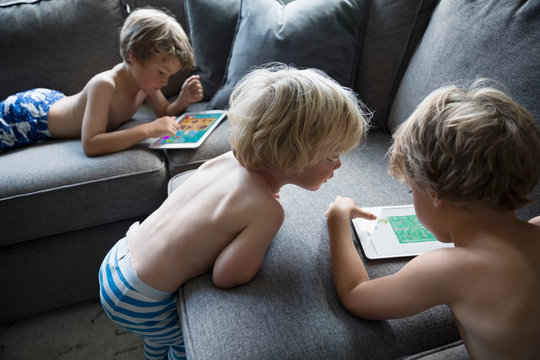 Bare chested boys in pajamas using digital tablets on living room sofa