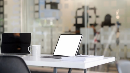 Cropped shot of workplace with blank screen tablet, laptop, office supplies and coffee cup on white table with blurred office room