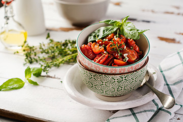 Baked cherry tomato with garlic, thyme and basil in vintage ceramic bowls on a light wooden table. Top view.