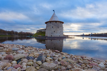 The watchtower of the Pskov Kremlin reflected in water.  In the foreground are stones of different sizes.