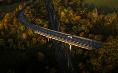 Winding road in autumn forest at sunset in mountains. Aerial view. Top view of beautiful asphalt roadway and orange trees. Highway through the woodland in fall. Trip in europe. Travel and nature - obrazy, fototapety, plakaty