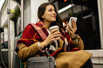 Portrait of a young woman at the public transport, sitting with coffee and using her cell phone  in...