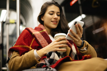 Portrait of a young woman at the public transport, sitting with coffee and using her cell phone  in...