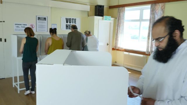 Asian Man voting at Polling Booth in the Election. Someone behind posts their vote into the Ballot Box. Polling place / Station.