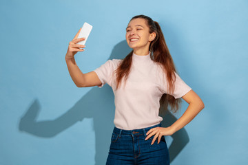 Taking selfie or vlog, smiling, posing. Caucasian teen girl's portrait on blue background. Beautiful model in casual wear. Concept of human emotions, facial expression, sales, ad. Copyspace. Looks