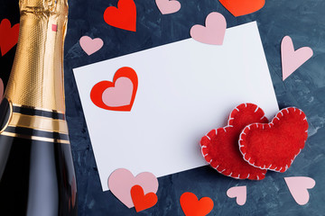 Valentine's day greeting card with two handmade hearts made of red fabric, bottle of champagne and several scattered hearts of various shapes cut out of paper on  textured background with copy space