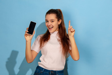 Showing phone screen, pointing up. Caucasian teen girl's portrait on blue background. Beautiful model in casual wear. Concept of human emotions, facial expression, sales, ad. Copyspace. Looks happy.