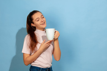 Enjoying coffee, tea, looks calm. Caucasian teen girl's portrait on blue background. Beautiful model in casual wear. Concept of human emotions, facial expression, sales, ad. Copyspace. Looks happy.