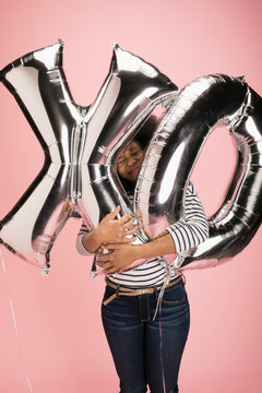 Young woman hugging silver XO balloons against pink background