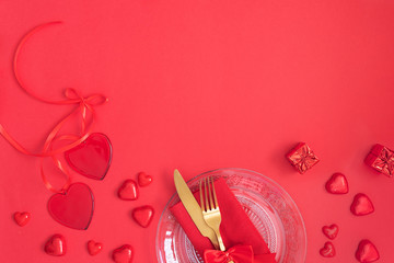 Festive table setting with Gold cutlery and crystal plate, red hearts and gift on red background.Saint Valentines day celebration or romantic dinner concept.