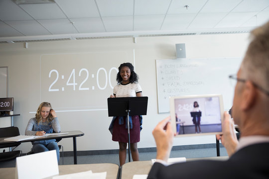 Teacher With Digital Tablet Videoing Girl Middle School Student Speaking In Debate Classroom