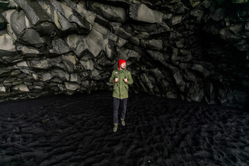 young male traveler in green clothes in a red hat and a green backpack in a black cave in Iceland