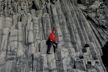 young male traveler in red clothes in a red hat and a green backpack stands on basalt pillars in...