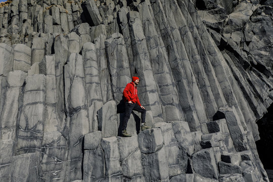 young male traveler in red clothes in a red hat and a green backpack stands on basalt pillars in Iceland