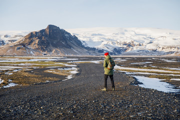 young male traveler in green clothes in a red hat and a green backpack walks along the path against the background of mountains in Iceland