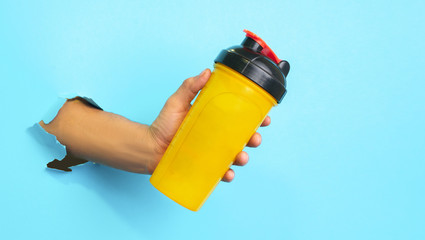 Man's fist holding yellow whey protein shake, ready to drink it, on blue background