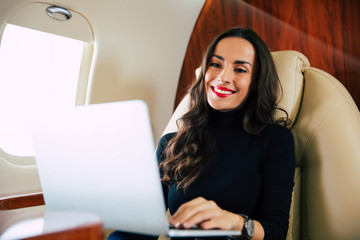 On top of the world. Close-up photo of an attractive girl with long chestnut hair, who is smiling, while typing something on her laptop during the flight.