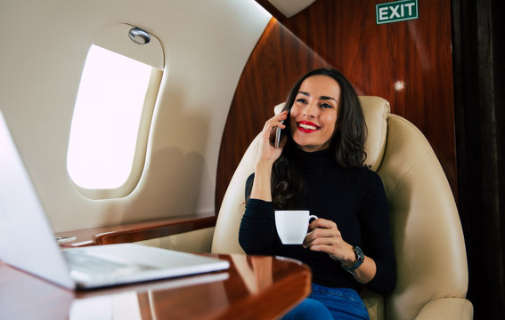 Talking with a friend. Close-up photo of a beautiful woman in a casual outfit, who is talking on the phone and drinking black coffee during her flight in private jet.