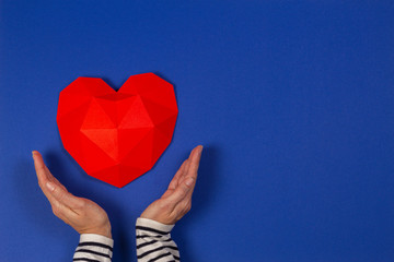 Female hands holding red polygonal heart on blue background. Top view