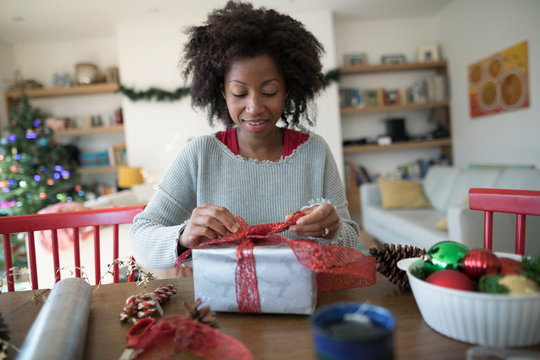 Woman wrapping Christmas gift at dining table, tying bow with red ribbon