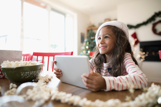 Smiling girl using digital tablet, wearing Christmas Santa hat and stringing popcorn decoration at table