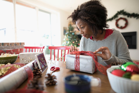 Woman watching video tutorial on digital tablet, wrapping Christmas gift at dining table