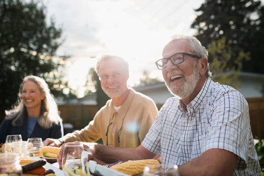 Laughing senior man enjoying garden party lunch with friends at sunny patio table