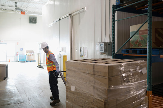 Worker pulling pallet jack with cardboard boxes in distribution warehouse