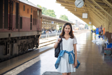 Portrait shot young Asian woman in white dress touching backpack on shoulder, lookink at camera and smile on face at train station, Travel and vacation concept.