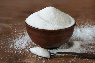 White sugar in a ceramic bowl of red clay and in a wooden spoon on a wooden background.