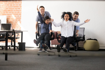 Excited diverse colleagues racing on chairs entertaining in office