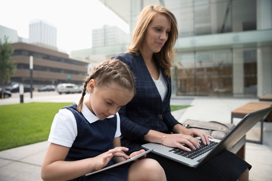 Businesswoman mother and schoolgirl daughter using laptop and digital tablet on urban bench