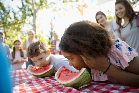 Boy And Girl Enjoying Watermelon Eating Contest At Summer Neighborhood Block Party In Park