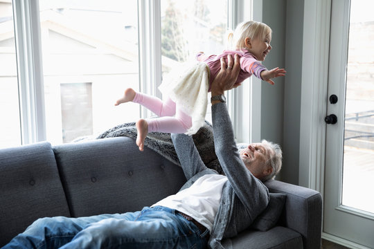 Playful grandfather lifting toddler granddaughter overhead on living room sofa