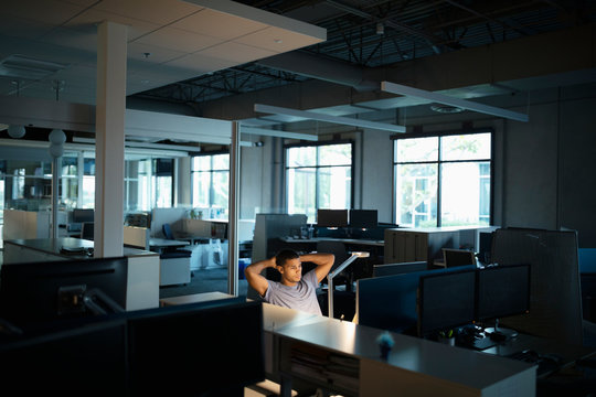 Businessman Working Late At Computer With Hands Behind Head In Dark Open Plan Office