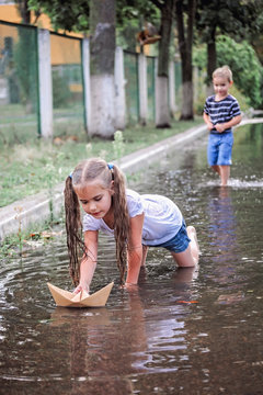 Cute happy kids playing with paper boat in the puddles after warm summer rain, lifestyle outdoor, back view