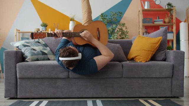 Joyful guy is playing the guitar singing wearing headphones having fun on sofa at home. Modern entertainment, music and happy young people concept.
