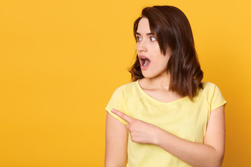 Close up portrait of young beautiful woman wearing casual t shirt, standing over isolated yellow background and points aside, looks worried and shocked. Copy space for advertisment or promotional text