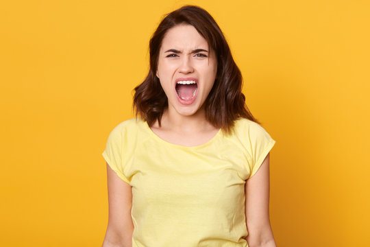 Horizontal shot of surprised young woman shouting angrily, posing isoalted over yellow background, adorable lady looking at camera woith angry facial expression, female wearing casual clothing.