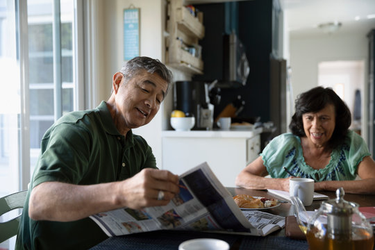 Latinx senior couple reading newspaper and eating breakfast at kitchen table