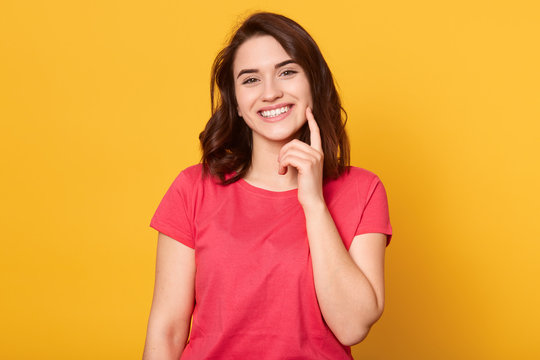 Cheerful young Caucasian woman with gentle charming smile, wearing red shirt, having pleasant appearance, keeps index finger on cheek, waits kiiss, smiles happily, poses against yellow background.