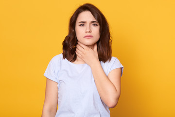 Close up portrait of cute sick young brunette woman wearing white casual t shirt, having sore throat, holding hand on her neck, sufferingfrom throat pain, painful swallowing, health care concept.