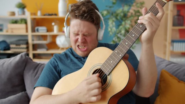 Creative guy with expressive face is playing the guitar at home wearing headphones having fun relaxing alone in apartment. People and emotions concept.