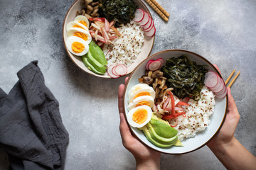 Poke bowl or sushi bowl with crab, rice, avocado, wakame seaweed, egg, radish and sesame seeds. Healthy clean eating. Top view, flat lay, gray background. Female hands holding Poke bowl