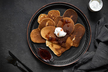 Heart shaped pancakes for Valentine's Day breakfast. Food with love. Pancakes with cream and strawberry jam. Top view, flat lay, dark background