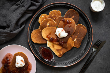 Heart shaped pancakes for Valentine's Day breakfast. Food with love. Pancakes with cream and strawberry jam. Top view, flat lay, dark background