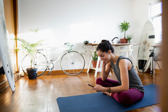 Young Latinx Woman With Smart Phone On Yoga Mat In Apartment