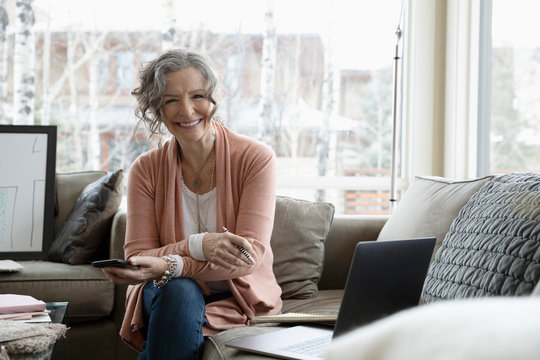 Portrait smiling, confident senior woman working from home, using smart phone and laptop on sofa